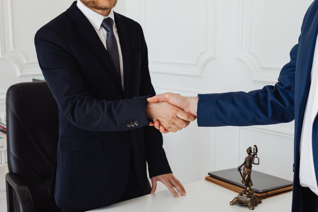 Home Two men in suits shaking hands in a formal office environment symbolizing an agreement.